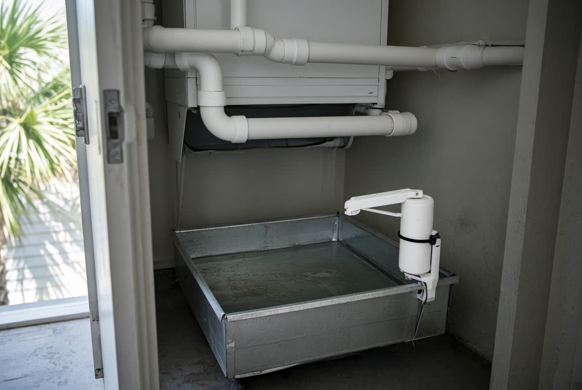 Coastal Florida air-handler closet with metal overflow pan holding water and a white float switch mounted high above the water line, PVC lines above, palm light through the open door